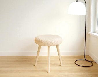 May include: A small, round, beige stool with a soft, textured seat and three pale wooden legs. The stool is placed on a light wood floor, with a floor lamp and a white wall in the background.
