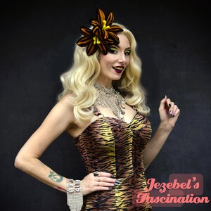 Peut inclure: Une femme blonde porte une robe &agrave; rayures de tigre et un accessoire de cheveux en forme de fleur. Elle porte &eacute;galement un collier &agrave; plusieurs rangs et un bracelet. L'image contient le texte "Jezebel's Fascination".