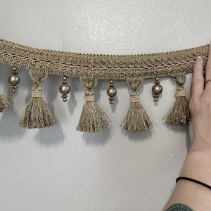 May include: Gold-toned decorative trim with tassels and beaded accents. The trim features a woven design with small tassels hanging below, each adorned with a round, pearl-like bead. The trim is likely used for embellishing curtains, clothing, or other craft projects.