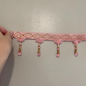 May include: Pink decorative trim with gold detailing and hanging beaded drops. The trim has a woven pattern with a scalloped border and small, teardrop-shaped beads. The trim is held against a neutral background.