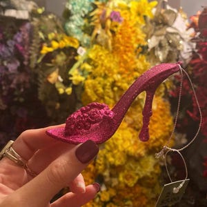 Könnte beinhalten: Ein funkelnder, fuchsiafarbener High-Heel-Schuh-Ornament mit floralem Design auf der Spitze. Der Stiletto-Absatz und der Schuh sind mit Glitzer bedeckt. Zum Aufhängen ist eine silberne Schnur angebracht.