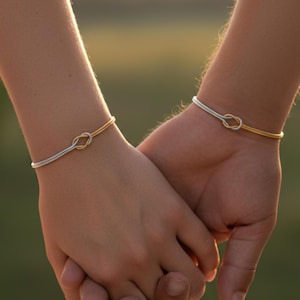 May include: Two hands clasped together, each adorned with a delicate bracelet featuring a knot motif. One bracelet is gold and silver, the other is silver. The background has a soft, blurred appearance with warm colours.