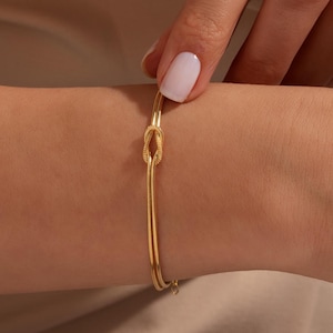 May include: A gold bracelet with a knot design at the centre. The bracelet has a delicate, chain-like appearance and is worn on a wrist. The background is a neutral colour, highlighting the jewellery.