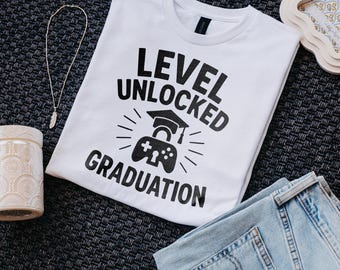 May include: White t-shirt with the text "LEVEL UNLOCKED GRADUATION" in black. The design includes a graduation cap and a video game controller. The shirt is paired with blue jeans and a silver necklace.