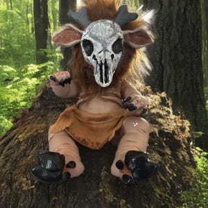 May include: A handcrafted fantasy creature doll with a skull mask, brown fur, and a tan loincloth. The doll has small gray antlers, large black feet, and is posed sitting on a tree stump in a forest setting.