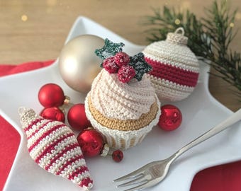 May include: A crocheted cupcake ornament topped with red berries and green leaves, accompanied by two crocheted ornaments with red and white stripes, several small red spherical ornaments, and a large champagne-colored sphere on a white star-shaped plate.