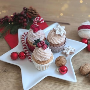May include: Three crocheted cupcakes on a white star-shaped plate. One cupcake features a red and white candy cane and red berries, another has a white snowflake, and the third has green holly leaves with red berries. Includes walnuts and red ornaments.