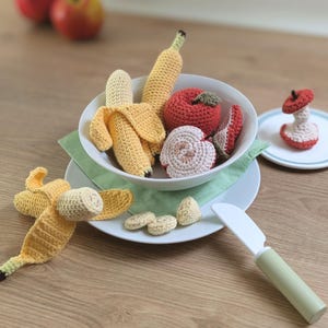 Peut inclure: Un ensemble de fruits en crochet comprenant des bananes jaunes, une pomme rouge, une pomme coupée et de petits morceaux de fruits dans un bol blanc. Un couteau jouet en plastique blanc avec un manche vert clair repose sur une assiette blanche.