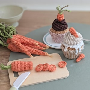Könnte beinhalten: Eine Sammlung von gehäkelten Spiellebensmitteln, darunter mehrere orangefarbene Karotten mit grünem Kraut, ein brauner Cupcake mit einer kleinen Karotte und ein beiger Cupcake mit weißem Zuckerguss und einer Karotte. Ein Holzbrett zeigt eine geschnittene Karotte.