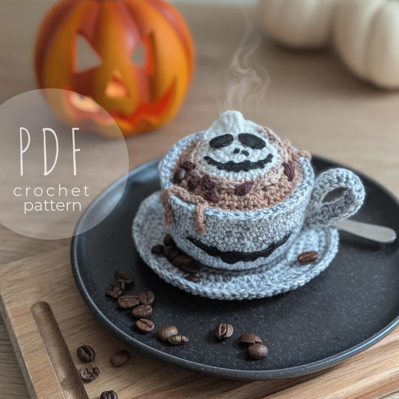 Peut inclure: Une tasse et une soucoupe &agrave; caf&eacute; crochet&eacute;es sur le th&egrave;me d'Halloween. La tasse est grise avec un visage de cr&acirc;ne souriant et remplie de caf&eacute; et de grains de caf&eacute; crochet&eacute;s bruns. La soucoupe contient des grains de caf&eacute;. L'image comprend une citrouille et le texte "PDF crochet pattern."