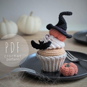 Peut inclure: Un cupcake d'Halloween crochet&eacute; avec un chapeau de sorci&egrave;re, une chauve-souris et une citrouille sur une assiette noire. Le cupcake a une base beige, un gla&ccedil;age blanc et une couche orange. Le chapeau de sorci&egrave;re est noir avec une bande violette. Le texte "PDF crochet pattern" est visible.