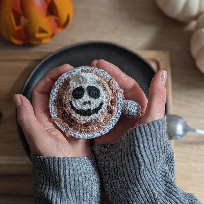 Peut inclure: Un dessous de verre d'Halloween crochet&eacute; avec un motif de cr&acirc;ne. Le dessous de verre est fait de fil beige, marron et gris. Le cr&acirc;ne a des yeux noirs et une bouche souriante. Une petite poign&eacute;e est attach&eacute;e sur le c&ocirc;t&eacute;.