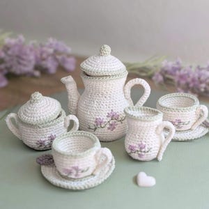 Può includere: Un set da tè all'uncinetto in filato color crema con bordi verde salvia e accenti floreali viola ricamati. Il set comprende una teiera con coperchio, una zuccheriera con coperchio, una lattiera e due tazze con piattini coordinati.