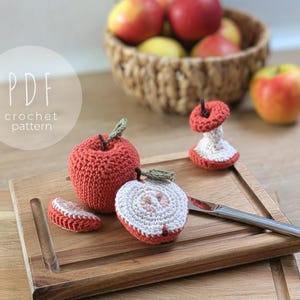 Op de afbeelding: Gehaakte items met appelthema, waaronder hele appels, appelschijfjes en een appelklokhuis, worden getoond op een houten snijplank. De appels zijn rood met groene bladeren, de kern is wit en rood. De tekst "PDF crochet pattern" is zichtbaar.