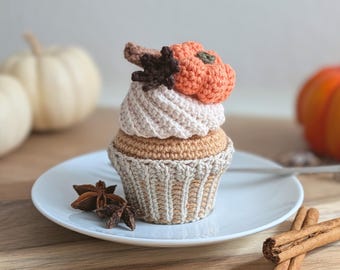 May include: A crocheted cupcake decoration featuring a tan base, a cream-colored swirled top, and a small orange pumpkin with a dark brown leaf accent. The item sits on a white plate alongside a star anise and two cinnamon sticks.
