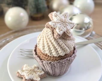 May include: A crocheted cupcake ornament featuring a brown base, a cream-colored swirled top, and a white star-shaped topper with gold embroidery. A separate small white star-shaped cookie with gold stitching sits on the white plate.