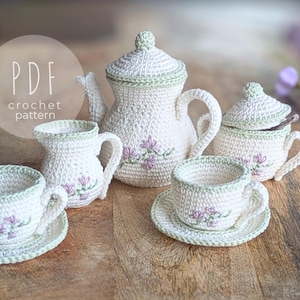 以下が含まれることがあります： クリーム色の、薄緑色の縁取りと紫色の花の刺繍が施されたかぎ針編みのティーセット。 ティーポット、クリーマー、砂糖入れ、ソーサー付きのティーカップ2客が含まれています。 「PDF crochet pattern」の文字が見えます。