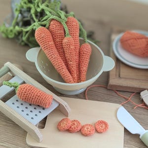 May include: A collection of crocheted orange carrots with green tops placed inside a light-coloured colander. A single crocheted carrot rests on a wooden grater, while four small crocheted carrot slices are arranged on a wooden cutting board.