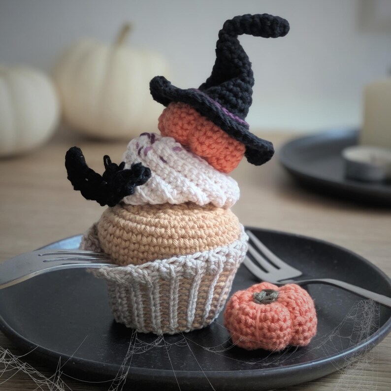 Peut inclure: Un cupcake d'Halloween crochet&eacute; avec un chapeau de sorci&egrave;re noir, une citrouille orange et une chauve-souris noire. Le cupcake est beige, blanc et orange, pos&eacute; sur une assiette noire avec une petite citrouille crochet&eacute;e.