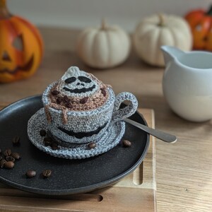 Peut inclure: Une tasse &agrave; caf&eacute; crochet&eacute;e sur le th&egrave;me d'Halloween avec une soucoupe, pr&eacute;sentant un motif de visage de fant&ocirc;me sur le caf&eacute; et la tasse. La tasse est grise avec un visage souriant noir. Des grains de caf&eacute; sont &eacute;parpill&eacute;s.