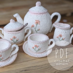 Op de afbeelding: Een gehaakte theeset in wit met roze randen en bloemenborduurwerk. De set bevat een theepot, suikerpot, melkkan en twee kopjes met schotels. De tekst "PDF crochet pattern" is zichtbaar.