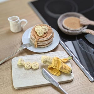 Puede incluir: Un conjunto de comida de juguete tejida a ganchillo que incluye una pila de tortitas con rodajas de plátano en un plato blanco, una jarra blanca pequeña, una sartén con una tortita, un plátano medio pelado y tres rodajas de plátano.