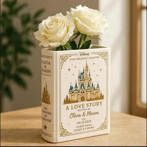 Op de afbeelding: Een rechthoekige keramische vaas in de vorm van een boek, voorzien van een gouden kasteelillustratie, de tekst Disney Our Greatest Chapter, A Love Story, Olivia & Mason en de datum 04.12.2025. Twee witte rozen staan in de vaas.