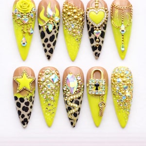 May include: A collection of ten long, pointed artificial nails. Each nail features a unique design with a yellow base, leopard print accents, and various embellishments like rhinestones, gold chains, and charms. The designs include stars, hearts, and floral elements.