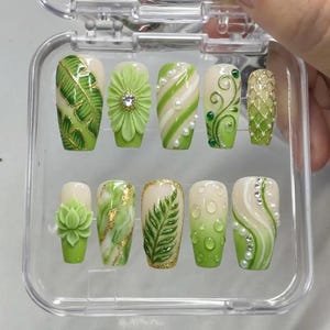 May include: A set of ten artificial nails in a clear plastic case. The nails feature various designs, including green leaves, floral patterns, and abstract swirls, with accents of gold glitter and small pearls. The color palette is primarily green and white.