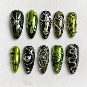 May include: A set of ten almond-shaped press-on nails in shades of olive and lime green with a metallic finish. The designs include silver 3D charms such as a star, cross, heart, spider web, snake, leopard print, and an eight-ball graphic.