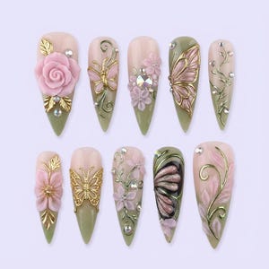 May include: A collection of ten artificial nails with various designs. Each nail features a light pink base with olive green tips. Embellishments include 3D flowers, butterflies, and gold accents. Some nails have rhinestones.