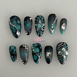 May include: A set of ten teal and black press-on nails with various designs. Some nails feature a marble effect, while others have zebra stripes or are adorned with rhinestones and silver star or flower accents. The nails are oval-shaped.