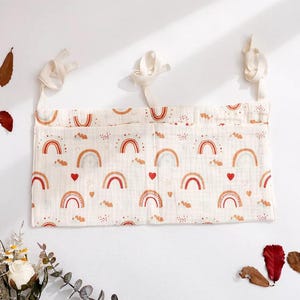 May include: A cream-coloured nursery organiser with a repeating pattern of rainbows, hearts, and clouds in shades of orange and red. The organiser has three pockets and is suspended by cream-coloured ties.