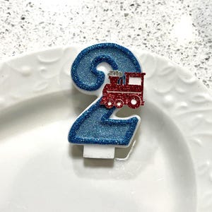 May include: A blue glitter number 2 birthday candle with a red train on top. The candle is white at the base and sits on a white plate. The candle is a festive decoration for a second birthday celebration.