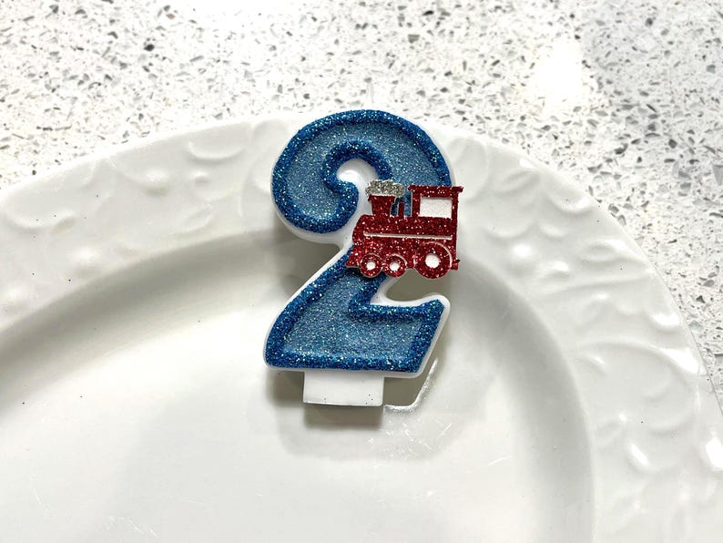May include: A blue glitter number 2 birthday candle with a red train on top. The candle is white at the base and sits on a white plate. The candle is a festive decoration for a second birthday celebration.