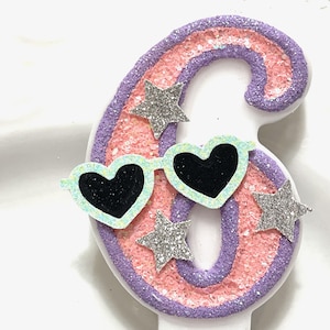 May include: A number six birthday candle with a pink and purple glitter design. The candle has heart-shaped sunglasses and silver glitter stars. The candle is white.