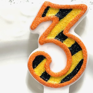 May include: A white number three candle with orange glitter edging and a yellow and black striped design. The candle is a festive decoration for a birthday or celebration.