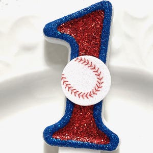 May include: A red and blue glitter birthday candle shaped like the number one with a white baseball on the front.