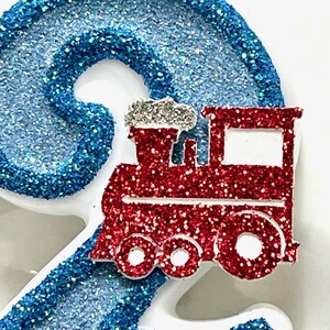 May include: A glittery blue number two with a red glitter train on top. The train has white accents and silver smoke. The number and train are likely cake toppers or decorations for a birthday.