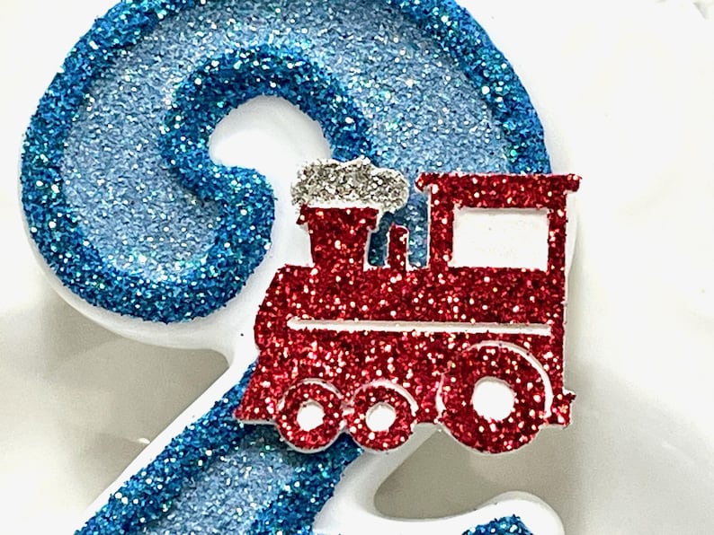 May include: A glittery blue number two with a red glitter train on top. The train has white accents and silver smoke. The number and train are likely cake toppers or decorations for a birthday.