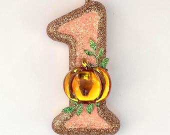 May include: A number one shaped birthday candle featuring a light pink center with a rose gold glitter border. The candle is decorated with a small, shiny orange pumpkin ornament and green glittered leaf accents attached to the front surface.