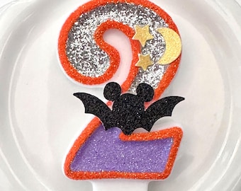 May include: A number two birthday candle featuring a silver glitter top section and a purple glitter bottom section, both outlined in orange glitter. A black glitter bat with mouse ears is attached to the center, with two gold stars and a crescent moon.
