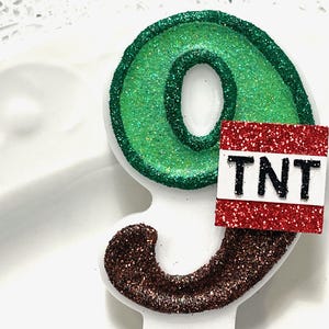 May include: Green number nine birthday candle with brown glitter around the bottom and a red glitter square with the word "TNT" in black letters.