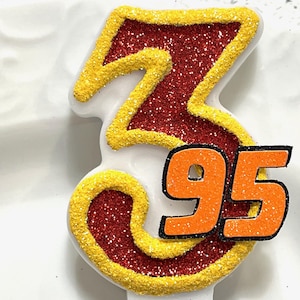 May include: Red and yellow glittery birthday candle shaped like the number 95. The number 95 is in orange with a black outline.