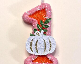 May include: A number one shaped birthday candle featuring a pink glittery border and a bright orange glittery center. The candle is decorated with a white glittery pumpkin cutout and a small green glittery leaf accent attached to the front.