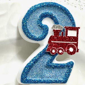 May include: A number two birthday candle with a blue glitter design and a red train. The number is outlined in blue glitter and has a white base. The train is red with silver accents.