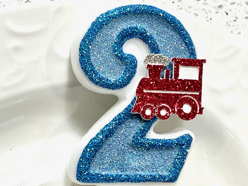 May include: A number two birthday candle with a blue glitter design and a red train. The number is outlined in blue glitter and has a white base. The train is red with silver accents.