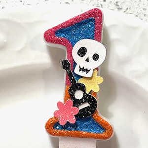 May include: A glittery orange, pink, and blue number one candle with a black and white skeleton playing a guitar. The skeleton has a pink flower on its head.
