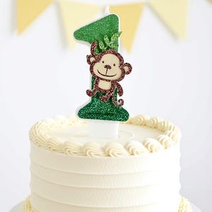 May include: A green glittery number one cake candle featuring a brown monkey illustration and small green leaves. The candle is inserted into the center of a round white cake with piped frosting borders along the top and bottom edges.