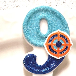 May include: A glittery number nine birthday candle. The number nine is light blue with a white base and a dark blue swirl. An orange and blue target design is attached to the number.